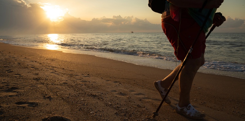 Image of an elderly person walking at sunrise with a walking stick on a beach