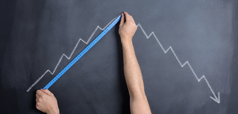 Image of hands holding a measuring tape to a jagged line trending up on a blackboard, while the line trends down beyond the measuring tape.
